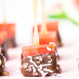 close up view of watermelon pops with wooden sticks covered in chocolate, coconut and sliced almonds on a pink tray