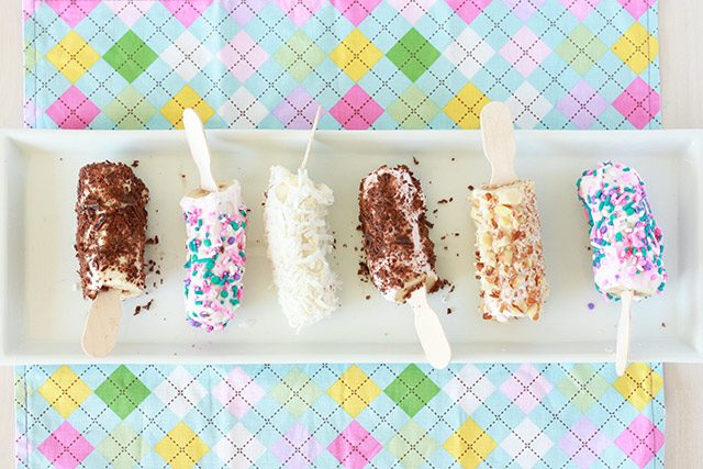 frozen banana pops on a white tray with different toppings: sprinkles, coconut, chocolate pieces, nuts