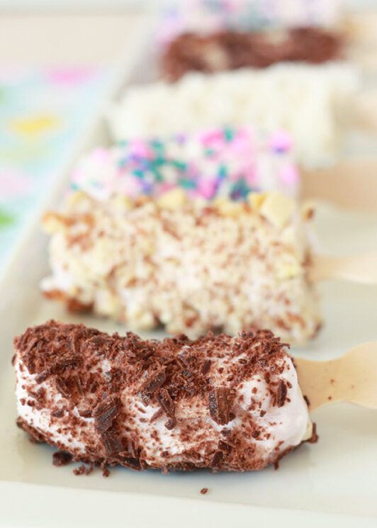 frozen banana pops on a white tray with different toppings: sprinkles, coconut, chocolate pieces
