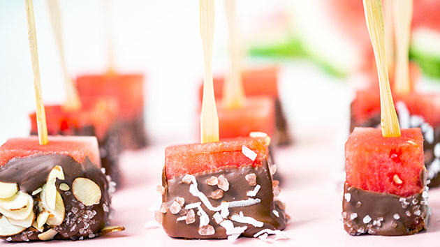 close up view of watermelon pops with wooden sticks covered in chocolate, coconut and sliced almonds on a pink tray