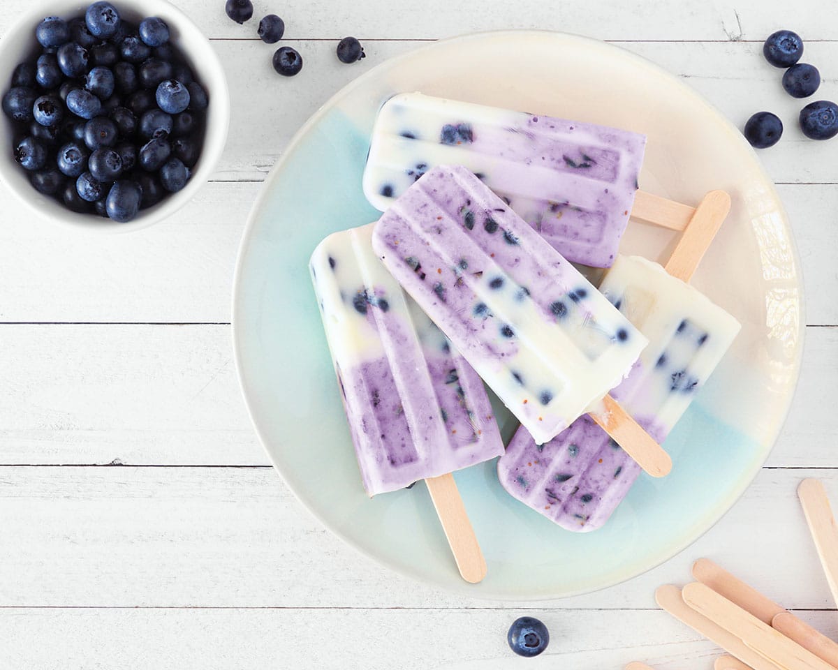 Plate of homemade blueberry vanilla yogurt popsicles. Top view corner border on a white wood background with copy space.