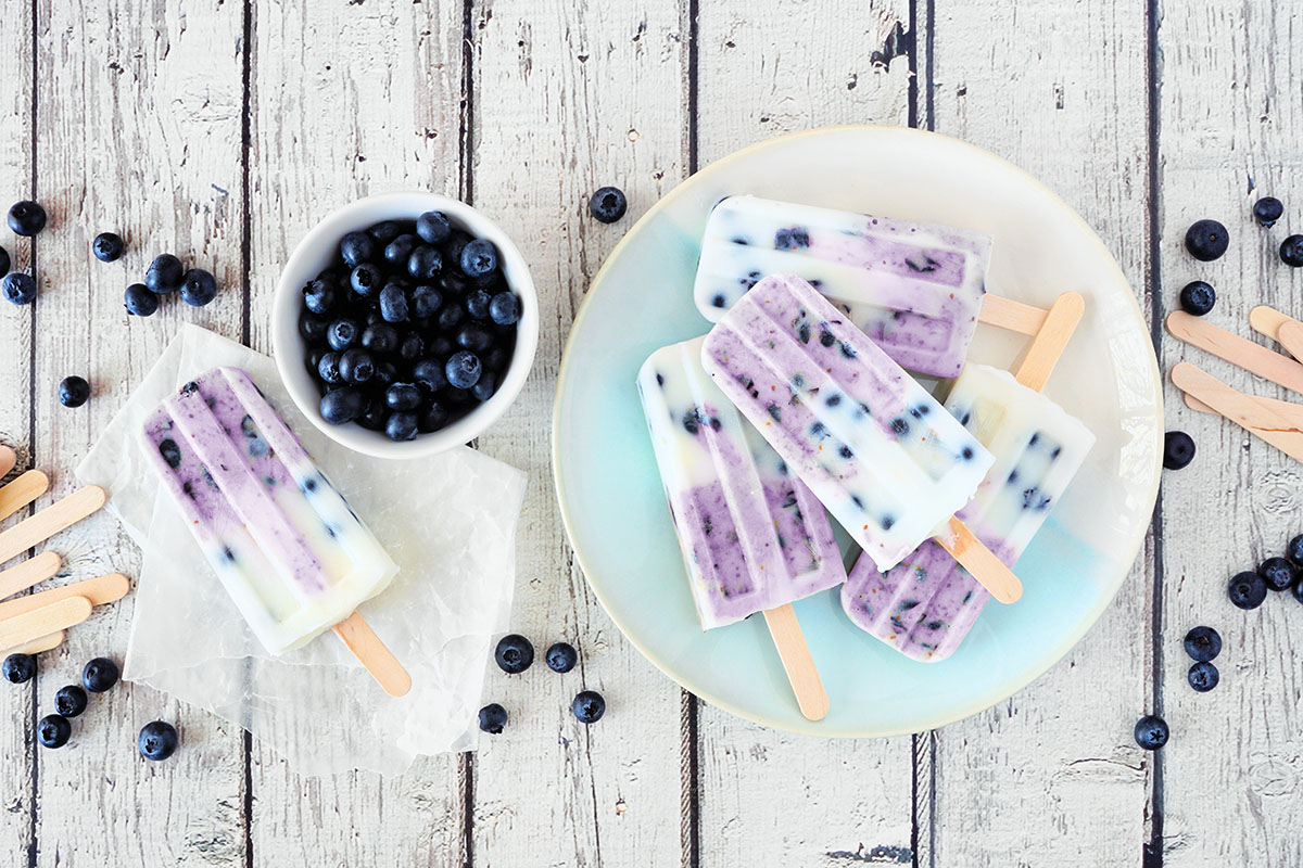 Blueberry vanilla yogurt ice pops. Top down table scene with a white wood background.