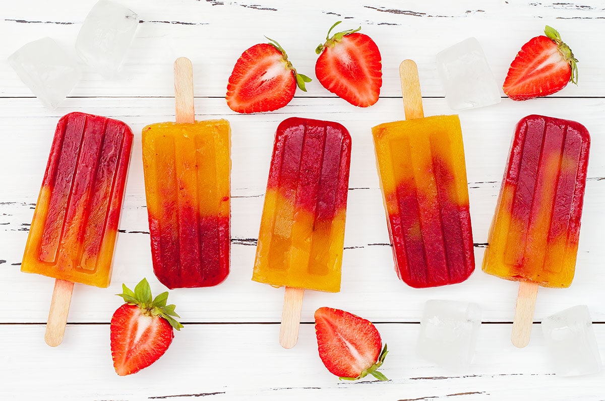 strawberry mango popsicles with fresh strawberries and ice cubes on a white board