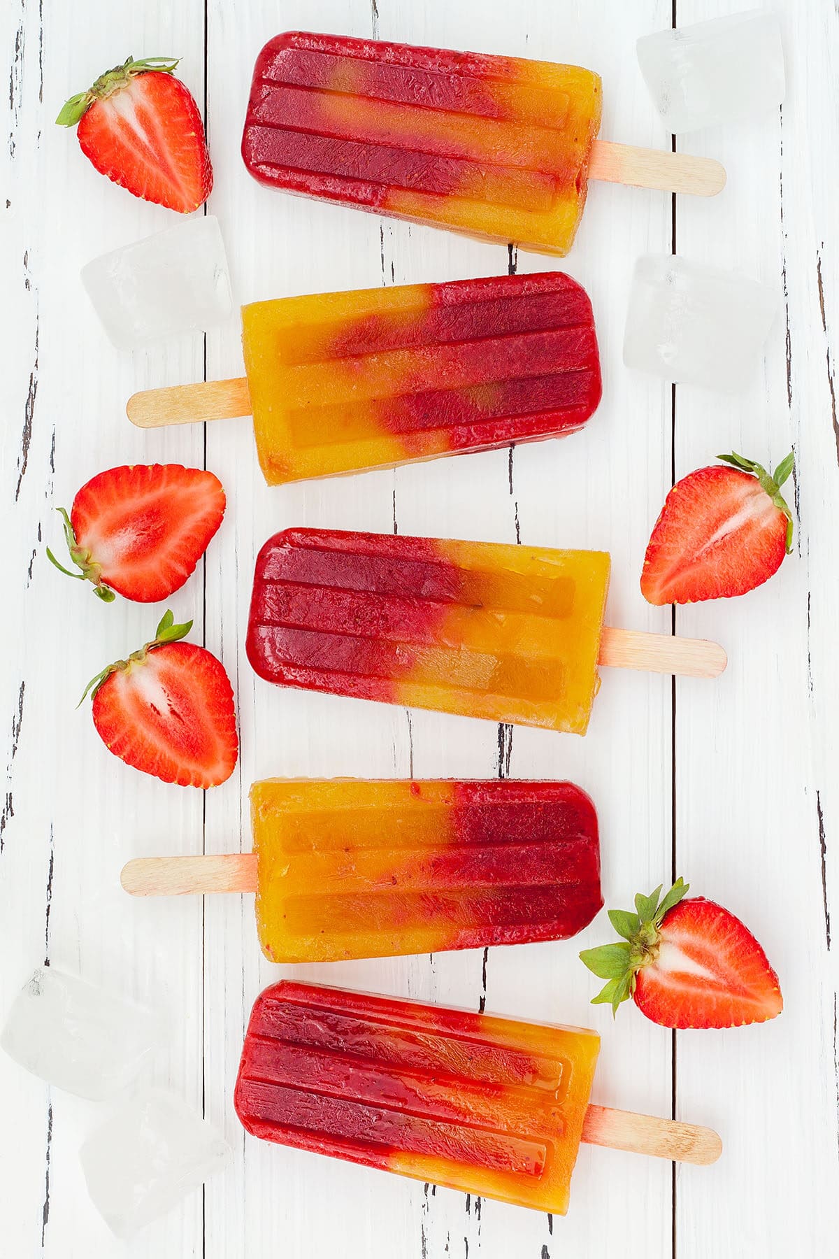 strawberry mango popsicles in a line with fresh strawberries and ice on a white board