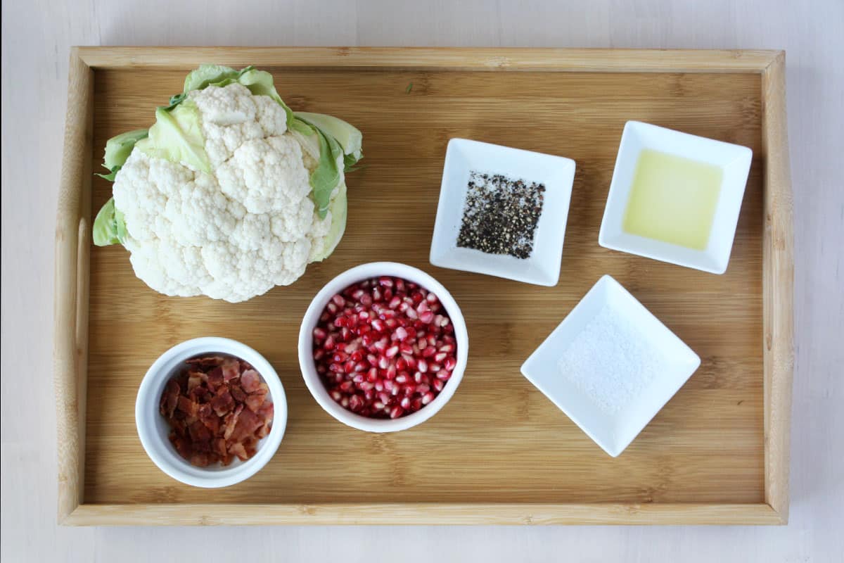 ingredients for roasted cauliflower - fresh head of cauliflower, bacon pieces, pomegranate arils, salt, pepper and oil.