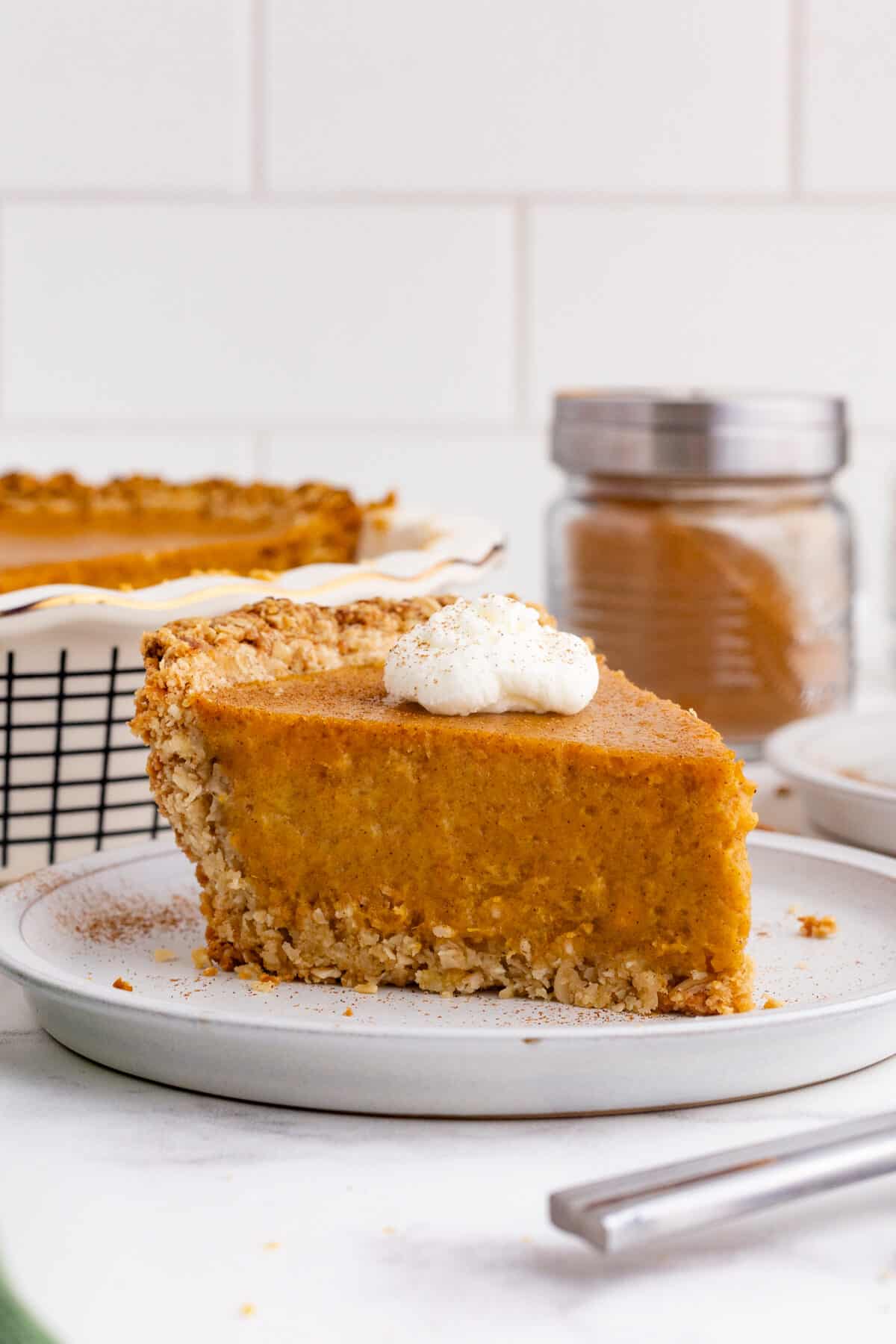 a slice of sweet potato pie on a plate with whip cream on top and the full pie and jar of cinnamon in the background