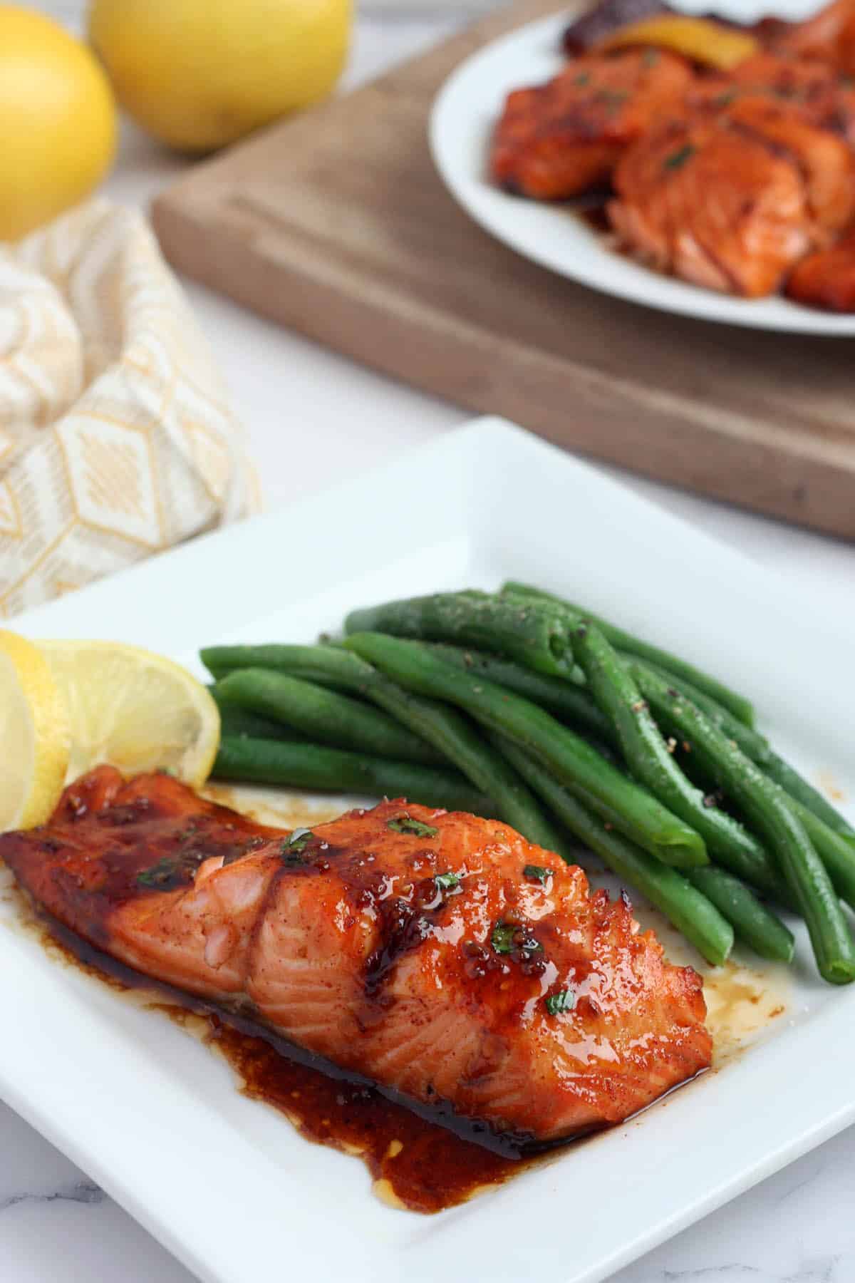 a fillet of honey garlic salmon on a white plate with green beans on the side and a lemon wedge