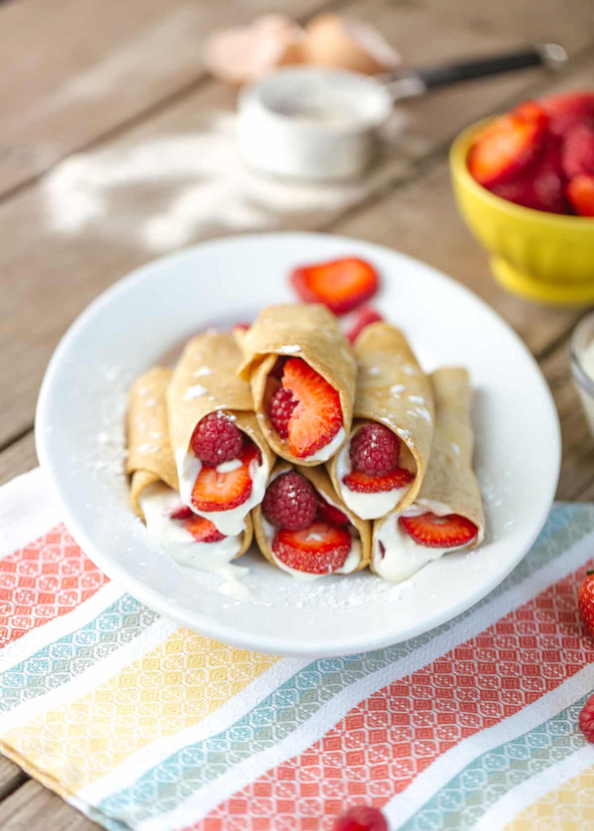 a plate of crepes filled with strawberries, raspberries and a cream filling on a white plate with a colorful napkin underneath