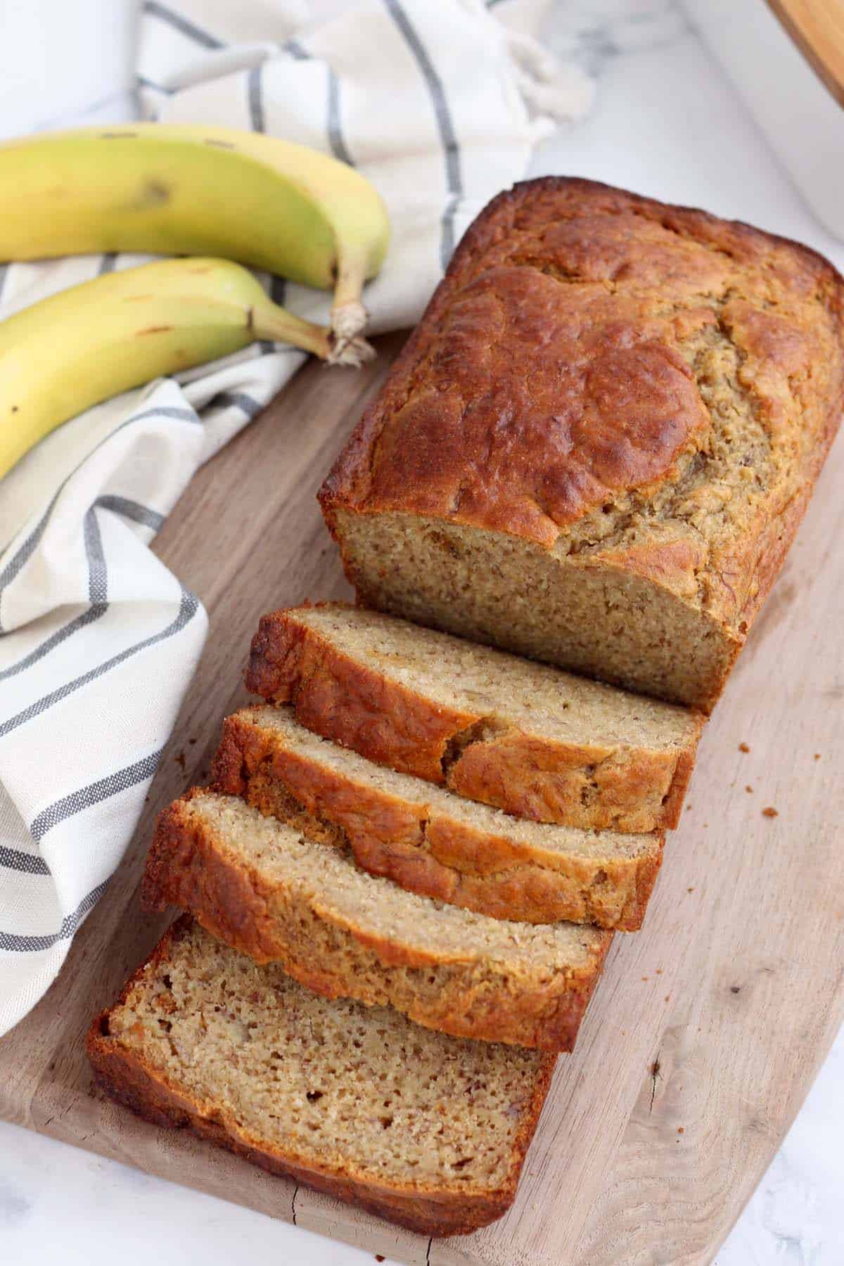 a loaf of healthy banana bread on a cutting board, half sliced with bananas in the background