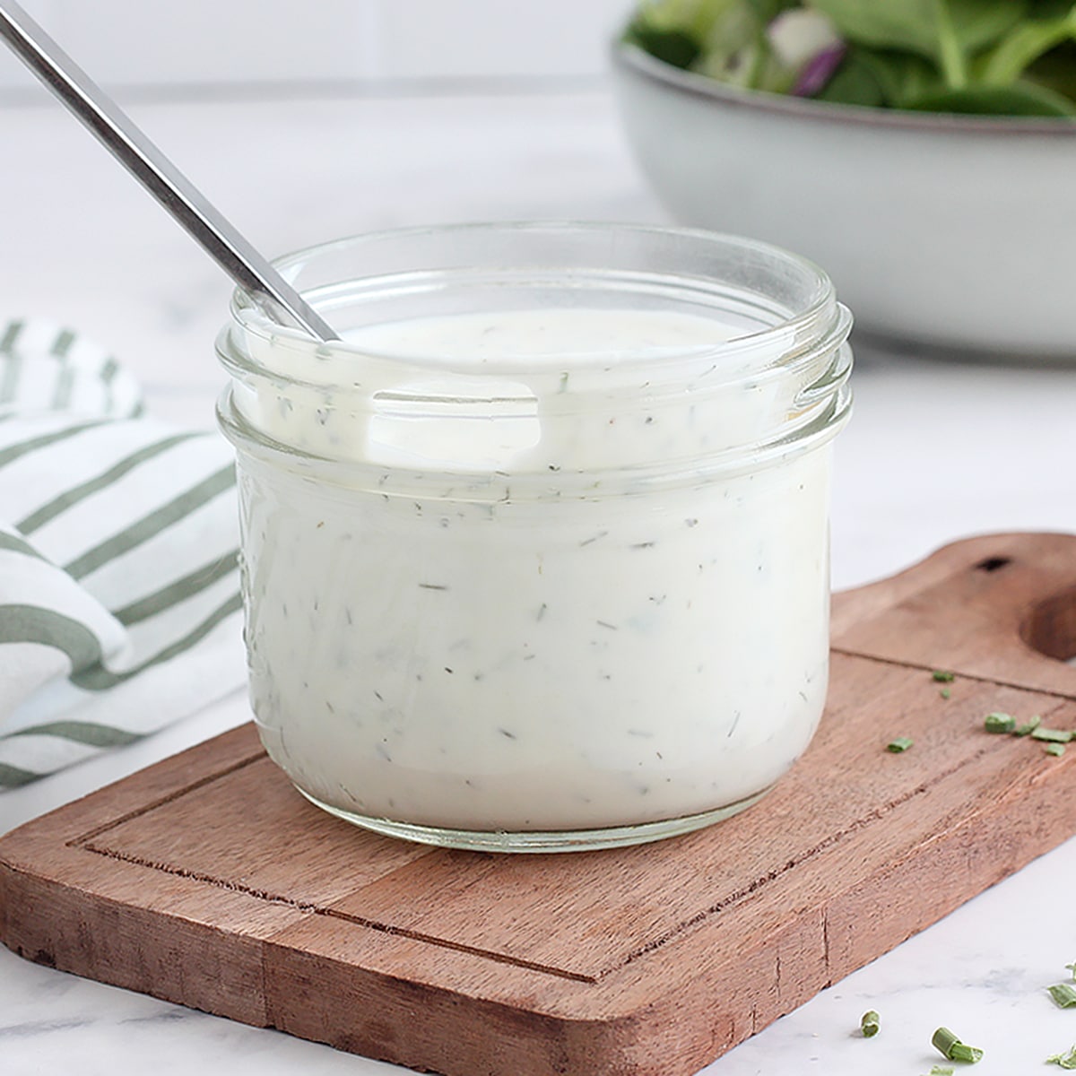 A small glass jar of ranch dressing with a striped linen and a metal spoon.