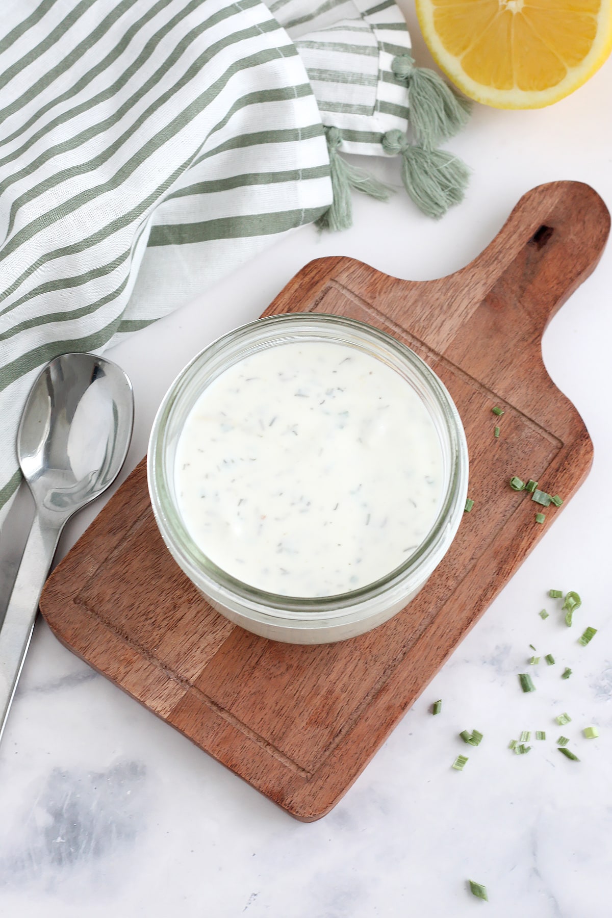 A jar of homemade ranch dressing on a small wooden board with a scattering of dried chives.
