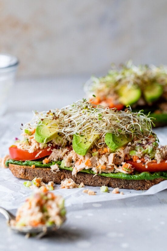 An open-faced tuna salad sandwich with avocado, with a forkful of canned tuna in the foreground.
