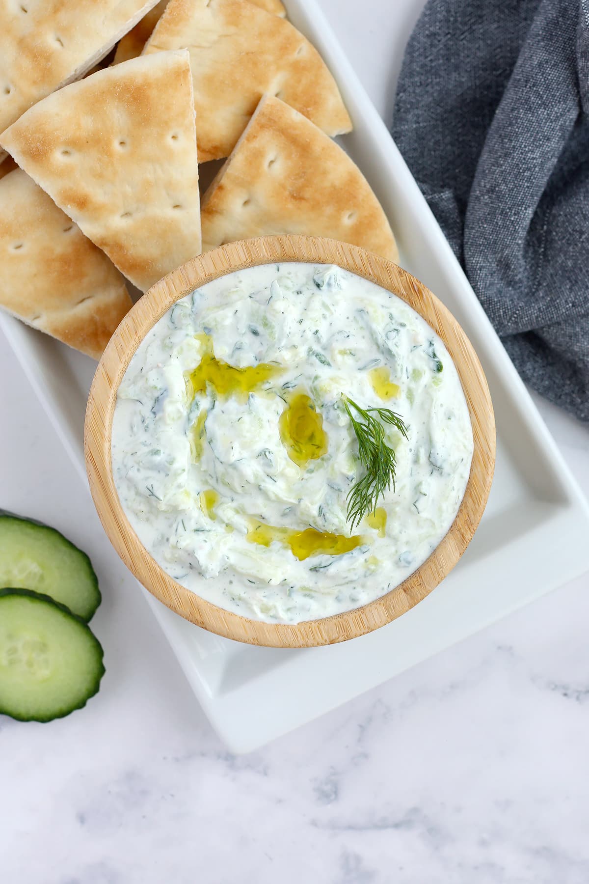 Homemade Tzatziki drizzled with olive oil on a white platter with pita bread and cucumber slices.