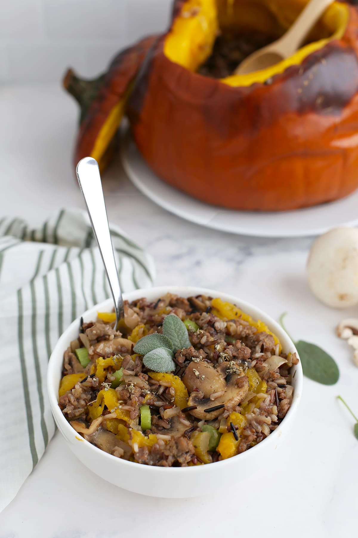 A bowl of dinner in a pumpkin with a silver spoon and topped with fresh sage.