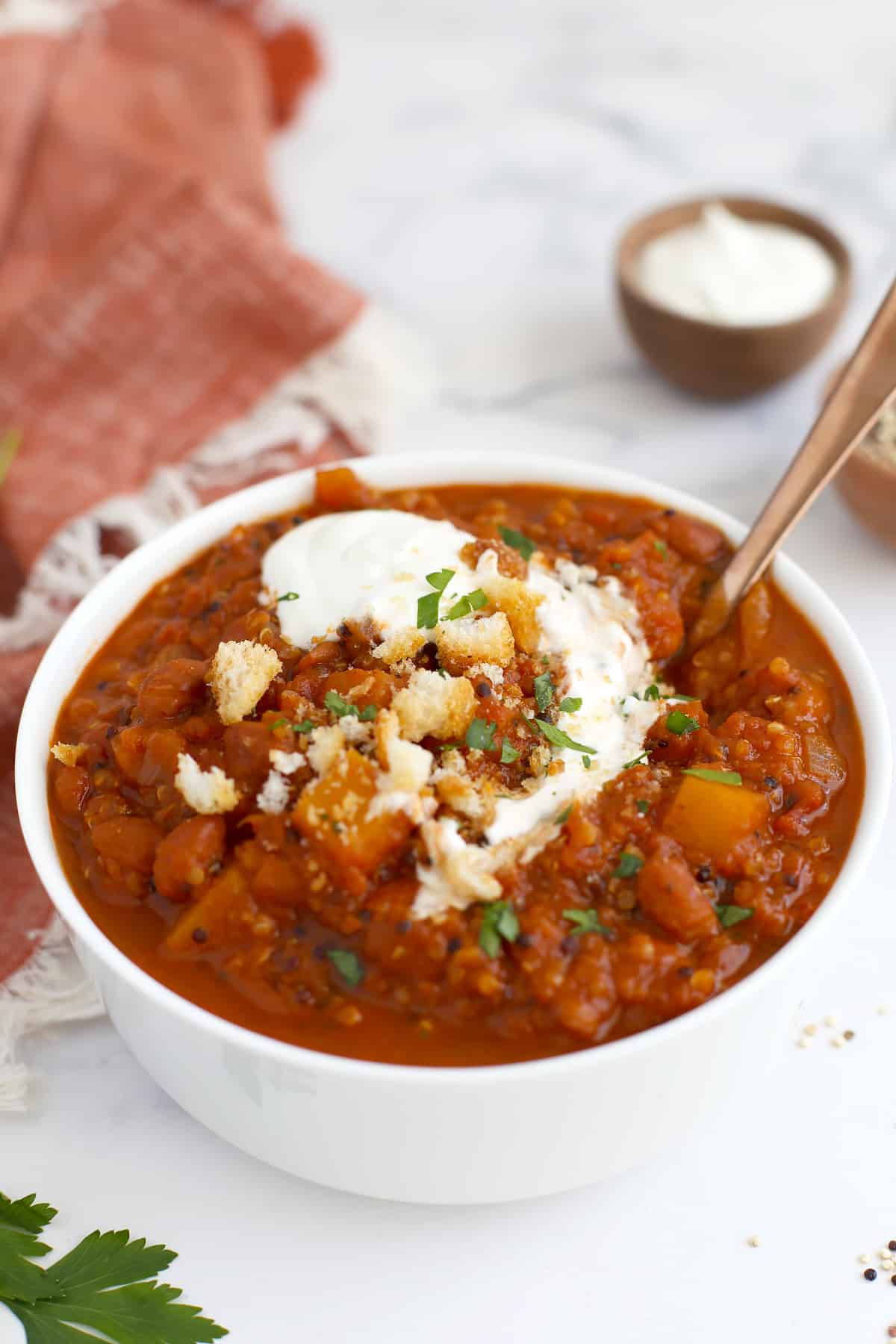A serving of pumpkin chili in a white bowl with a spoon, topped with fresh parsley and a swirl of sour cream.