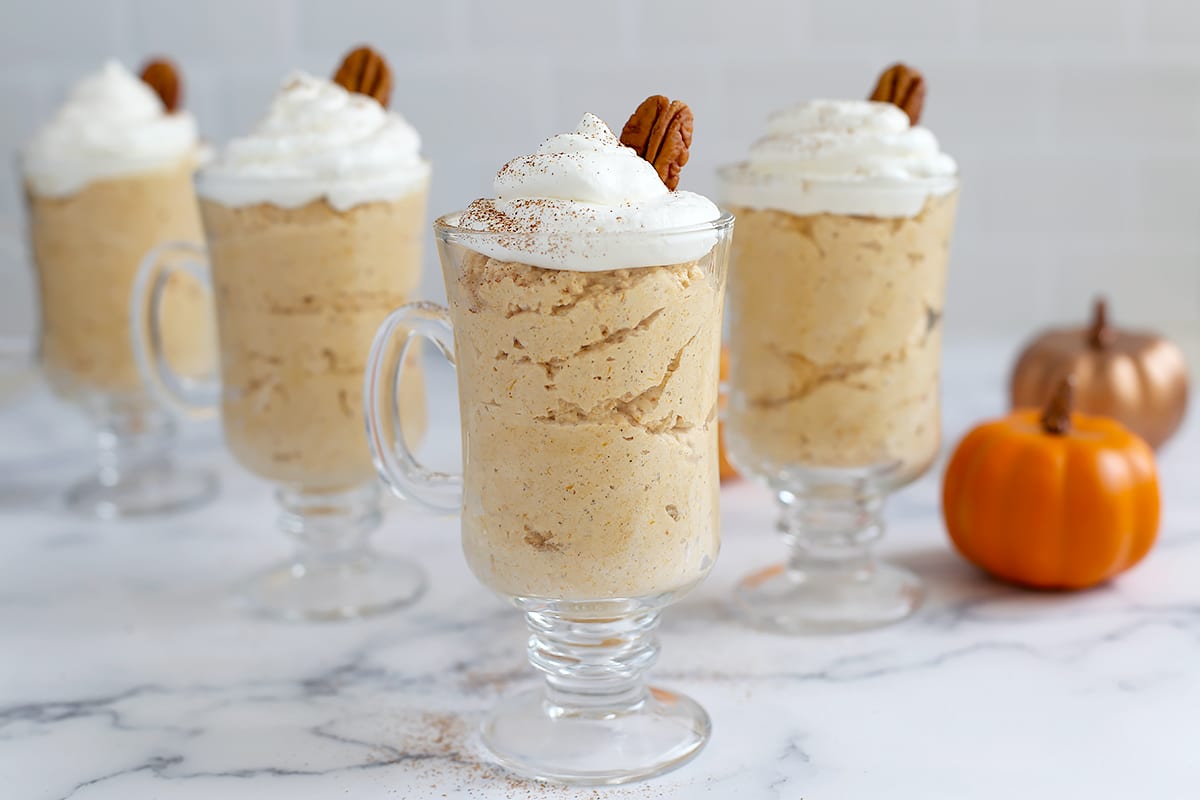 A row of pumpkin mousse desserts in glass serving jars topped with a swirl of whipped cream and a sprinkle of nutmeg.
