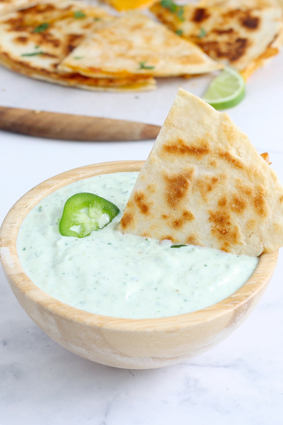 A triangle of quesadilla dunked in a bowl of cilantro ranch dressing topped with a slice of jalapeno.