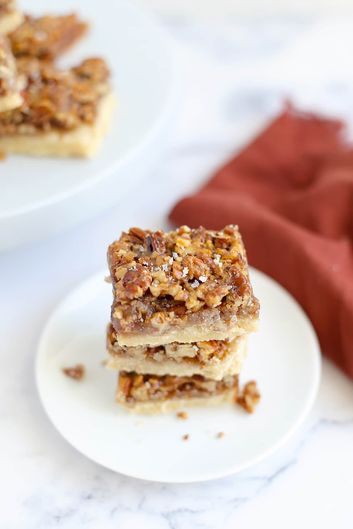 A stack of pecan pie bars sprinkled with flaked salt and stacked on a small serving plate with a red linen in the background.