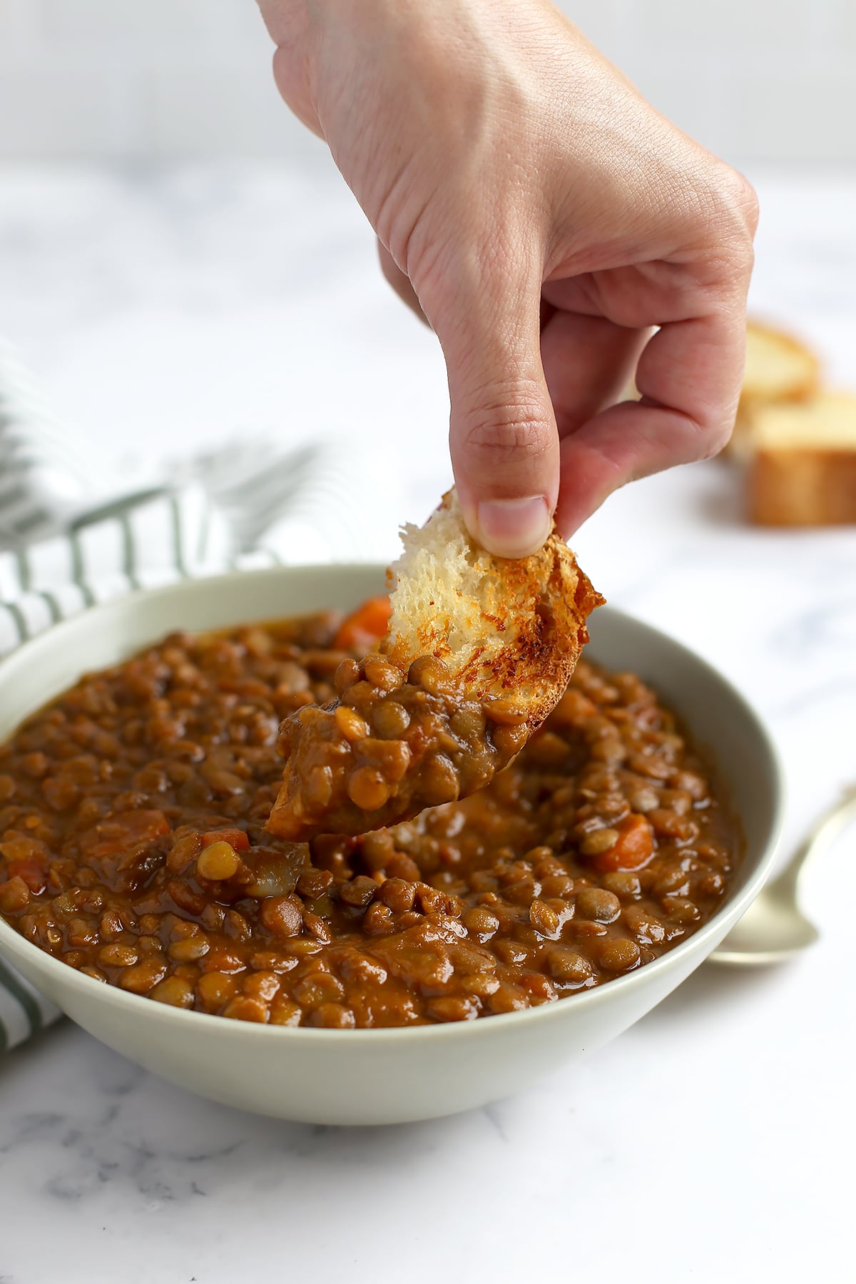 Someone dunking a crust of toasted bread into a bowl of hearty lentil soup.