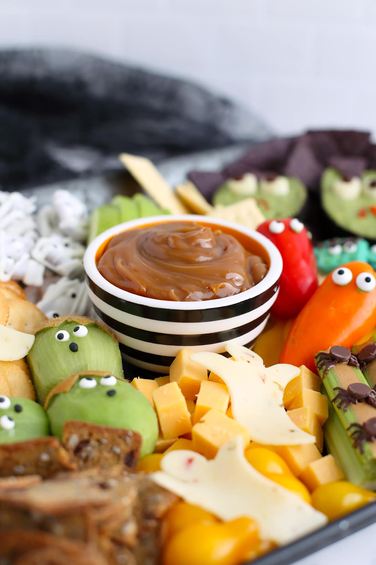 Caramel dip in a small striped bowl on a tray with cheese cubes, kiwi, chocolate covered pretzels, and assorted vegetables.