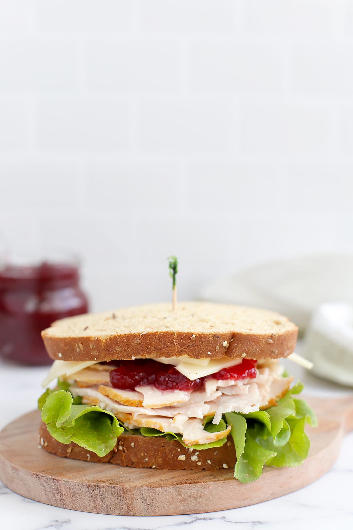 A turkey sandwich on wheat bread with cheese, cranberry sauce and lettuce on a wooden cutting board with a toothpick.
