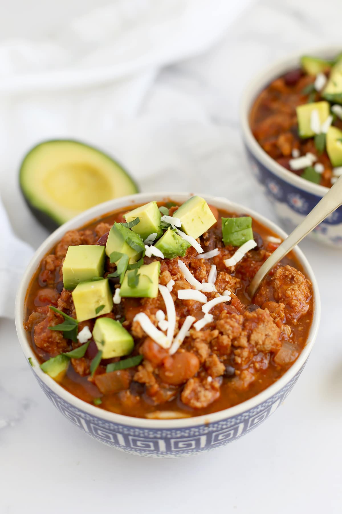 A close up image of homemade chili topped with avocado and cheese with a gold spoon.