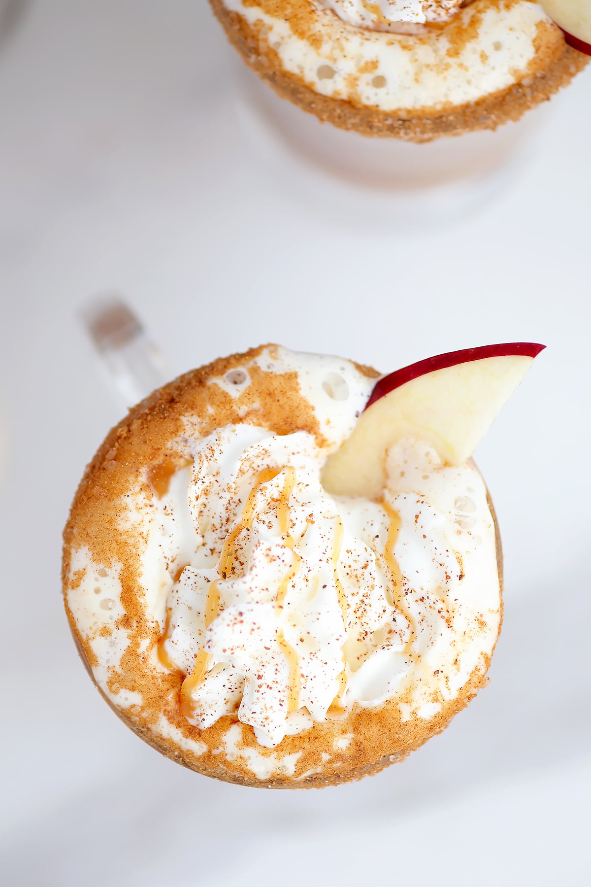 An overhead shot of a mug of cider topped with whipped cream, ground nutmeg, a caramel drizzle, and an apple slice.