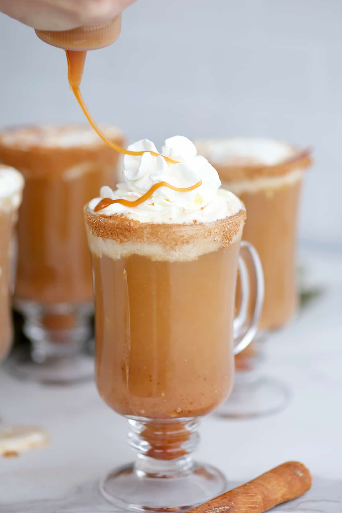 Homemade apple cider in a clear mug with whipped cream, a ribbon of caramel is being drizzled on top.