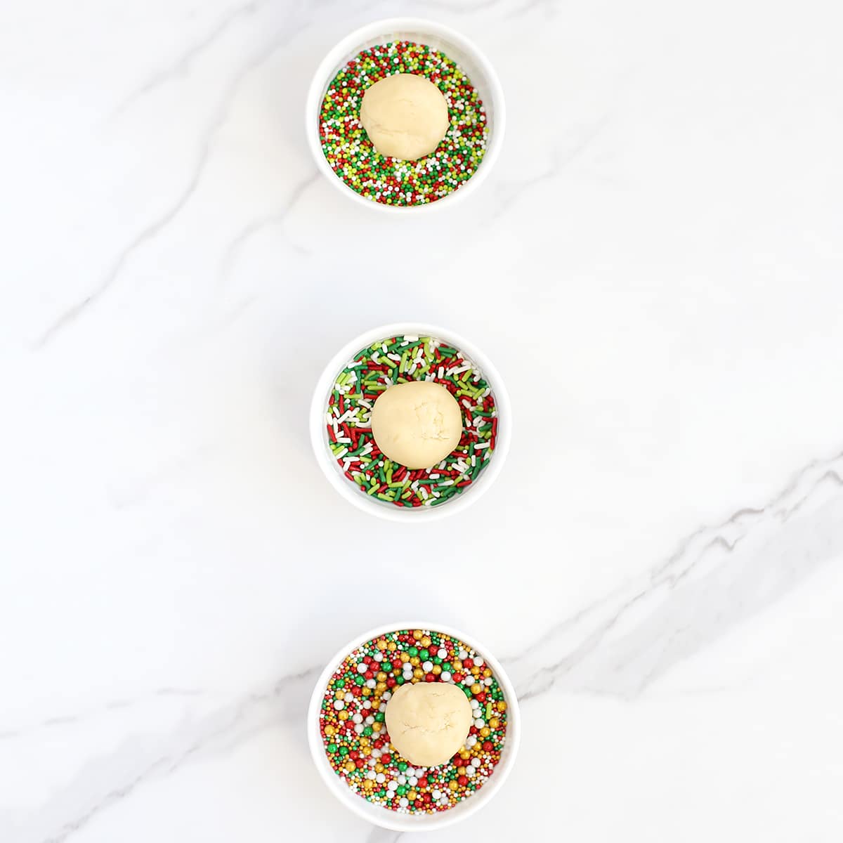 Three small bowls of sprinkles with a ball of cookie dough in each.
